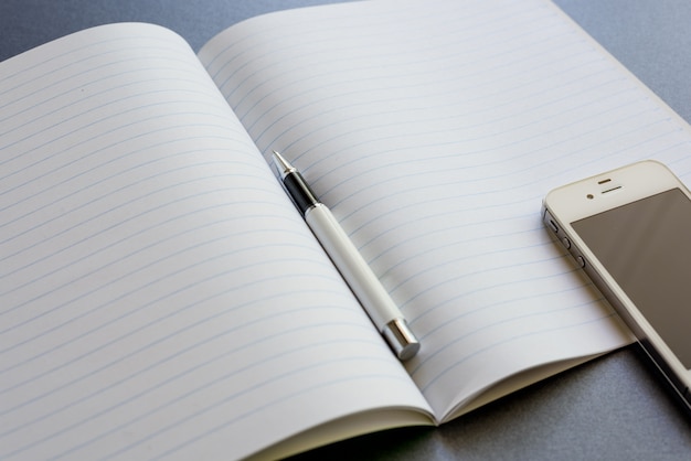 Un cahier ouvert avec un stylo et un téléphone mobile, sur un fond gris foncé, un travail de scène ou une étude.