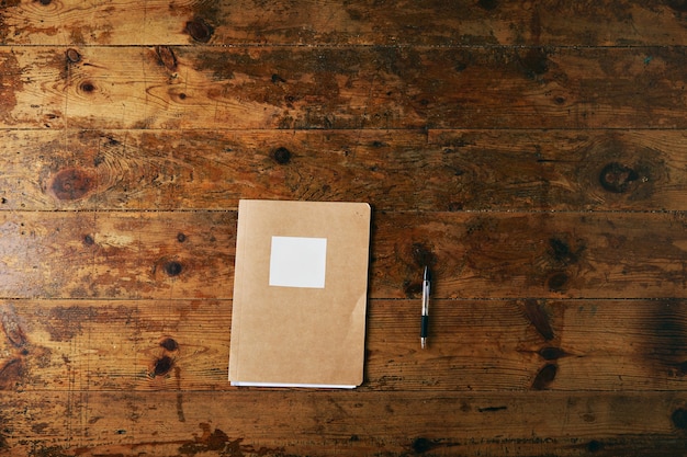 Cahier simple avec couverture en papier craft et étiquette blanche vide et un stylo à bille noir sur une table en bois brossé vieilli