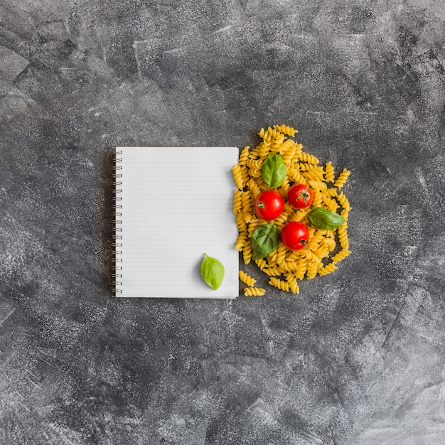 Cahier à spirale avec des fusilli crus; tomates et feuilles de basilic