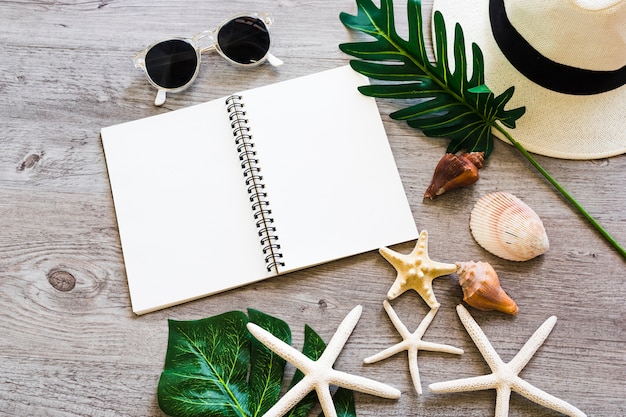 Photo gratuite cahier à spirale vierge avec coquillage, feuille, chapeau de paille, lunettes de soleil et étoile de mer sur table