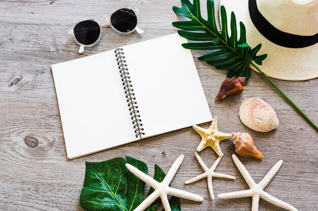 Cahier à spirale vierge avec coquillage, feuille, chapeau de paille, lunettes de soleil et étoile de mer sur table