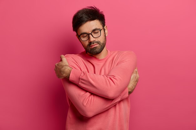 Calme bel homme barbu se câline, ressent le confort et le confort, se souvient d'une date romantique avec sa petite amie, ferme les yeux et incline la tête, porte un pull et des lunettes, isolé sur un mur rose