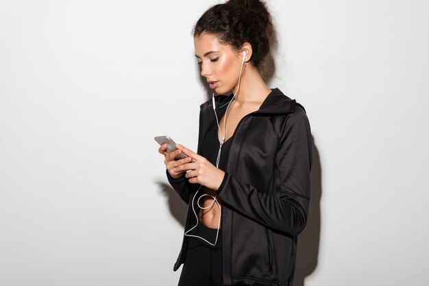 Calme femme brune bouclée de remise en forme, écouter de la musique et à l'aide de smartphone