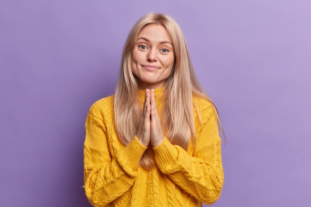 Calme heureux blonde jeune femme européenne presse les paumes ensemble et demande la faveur a besoin de votre aide porte un pull jaune décontracté