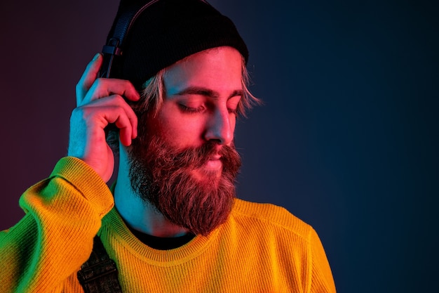 Calme, heureux. Portrait de l'homme caucasien sur fond de studio dégradé en néon. Beau modèle masculin avec un style hipster dans les écouteurs. Concept d'émotions humaines, expression faciale, ventes, publicité.