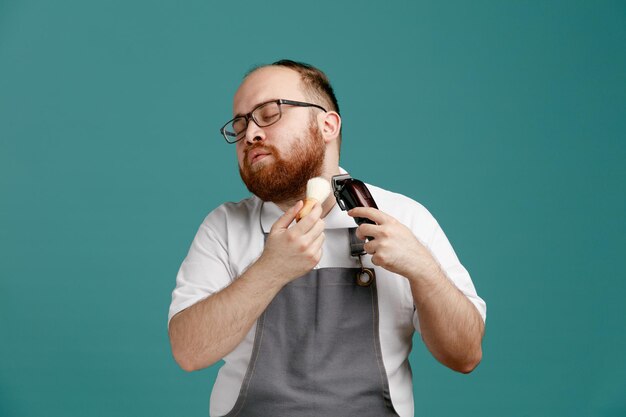 Calme jeune barbier portant l'uniforme et les lunettes tournant la tête vers le côté tenant le blaireau et la tondeuse à cheveux coupant le visage avec les yeux fermés isolés sur fond bleu