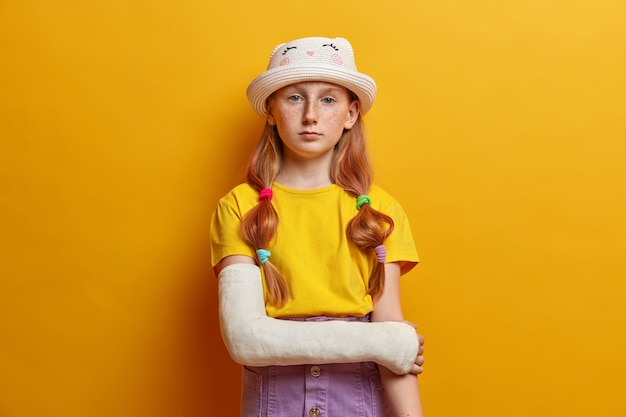 Calme petite fille sérieuse, a de longs cheveux roux et une peau tachetée de rousseur, porte une tenue d'été à la mode, pose avec la main dans le plâtre, récupère après un accident, isolé sur un mur jaune