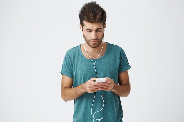 Camarade bronzé avec une coupe de cheveux élégante avec des amis sur un mur blanc. Jeune homme, porter, écouteurs, regarder, quoique, répondre, à, sien, petite amie, message, utilisation, wi-fi