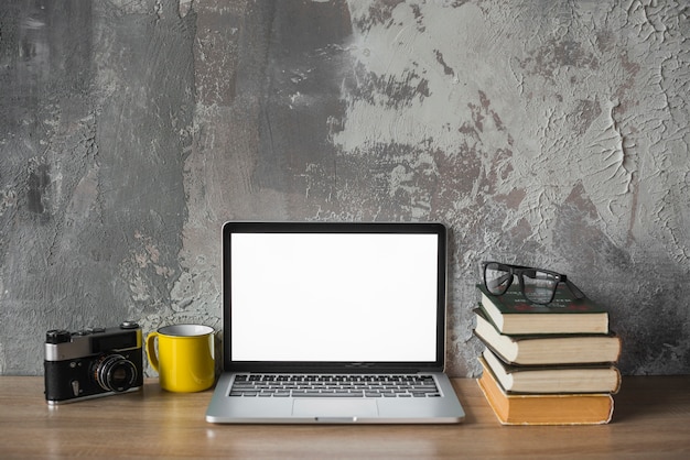 Caméra; tasse; livres empilés; spectacles et ordinateur portable avec un écran blanc sur un plateau en bois