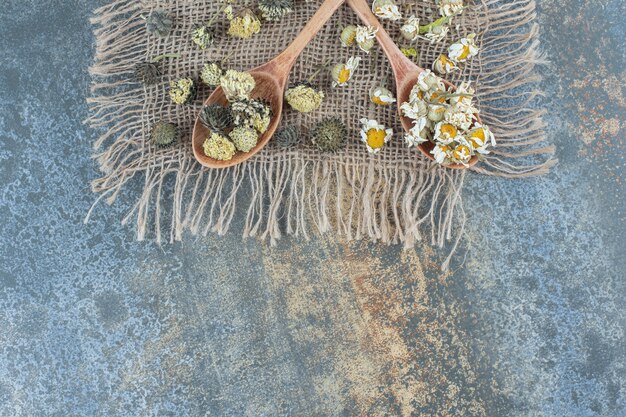 Photo gratuite camomille séchée et autres fleurs sur toile de jute avec cuillères en bois.