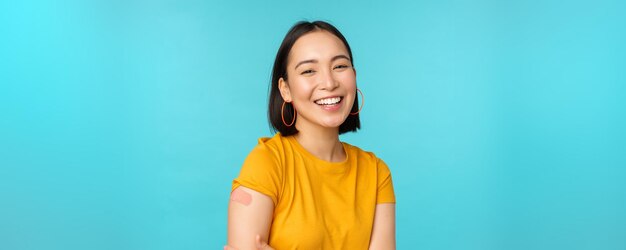 Campagne de vaccination contre covid19 Fille asiatique heureuse et en bonne santé riant après la vaccination contre le pansement contre le coronavirus sur l'épaule portant un t-shirt jaune sur fond bleu