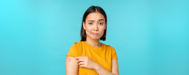 Campagne de vaccination de covid19 Femme asiatique triste et déçue montrant l'épaule avec un pansement et grimaçant contrarié debout sur fond bleu