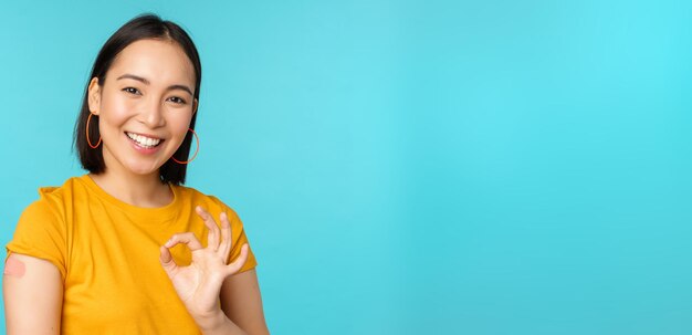 Campagne de vaccination de covid19 Heureuse fille asiatique en bonne santé montrant un signe correct et un pansement souriant sur l'épaule de la vaccination contre le coronavirus fond bleu