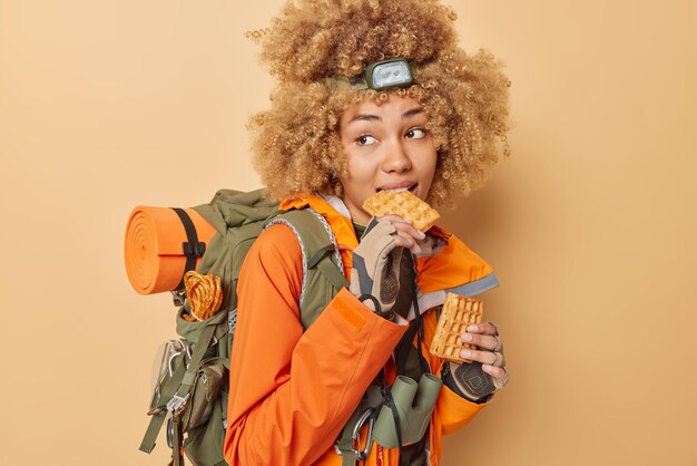 Une campeuse active aux cheveux bouclés et pensive a une délicieuse collation mange des gaufres maison se repose après la marche porte un sac à dos lourd porte une lampe frontale et une veste orange isolée sur fond beige