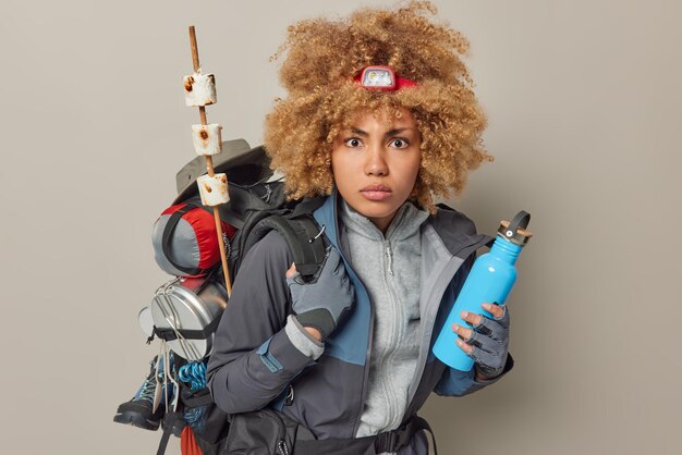 Une campeuse sérieuse revient d'un pique-nique regarde étonnamment avec un sac à dos contenant une bouteille d'eau fraîche vêtue de vêtements de sport isolés sur fond gris Temps d'aventure et loisirs