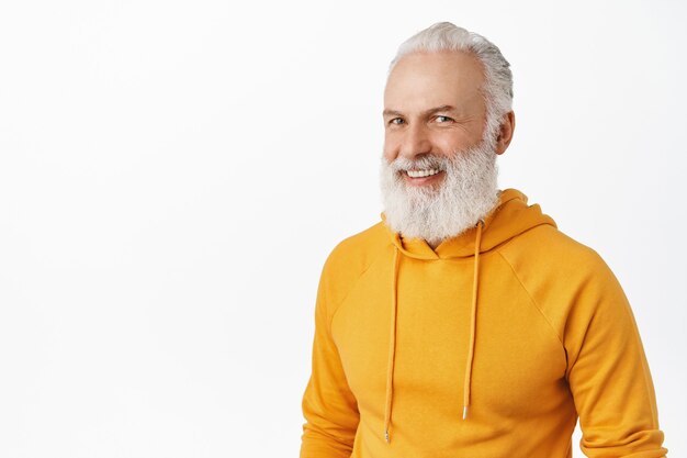 Candide vieil homme heureux avec une longue barbe élégante, riant et souriant, l'air heureux, portant un sweat à capuche orange moderne, debout sur un mur blanc