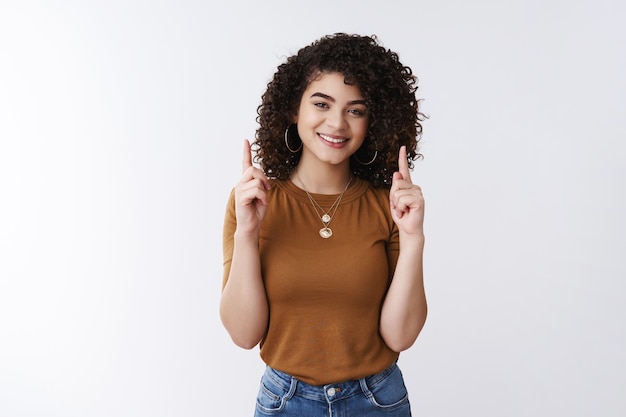 Cap en haut. Une jolie fille aux cheveux bouclés charismatique et sympathique lève l'index pointant vers le haut souriant gai montrant un graphique faisant la promotion du produit, debout publicité sur fond blanc