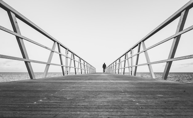 Capture en niveaux de gris d'une personne marchant sur un pont en bois au bord de l'océan - concept : départ, séparation