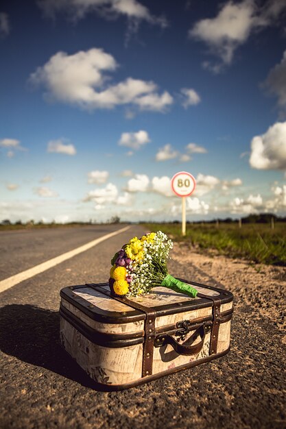 Capture verticale d'une valise vintage avec un bouquet sur une route vide