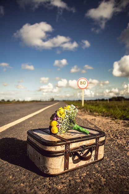 Capture verticale d'une valise vintage avec un bouquet sur une route vide