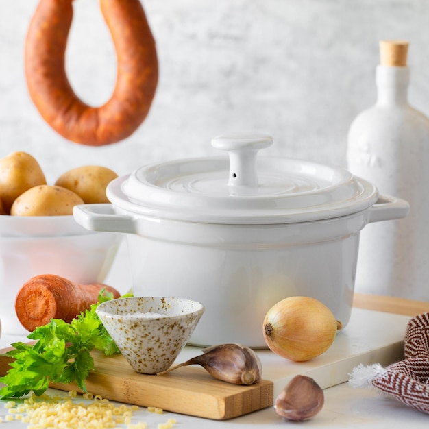 Une casserole blanche et des ingrédients pour faire de la soupe Minestrone avec des saucisses fumées sur une table blanche Soupe italienne traditionnelle avec des pâtes et des légumes de saison