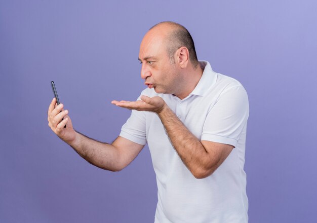 Casual homme d'affaires mature tenant et regardant le téléphone mobile et envoi de baiser coup isolé sur fond violet