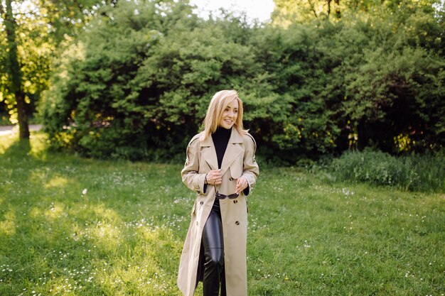 Caucasian blonde woman wearind trench sourire joyeusement le jour de printemps ensoleillé à l'extérieur à pied dans le parc