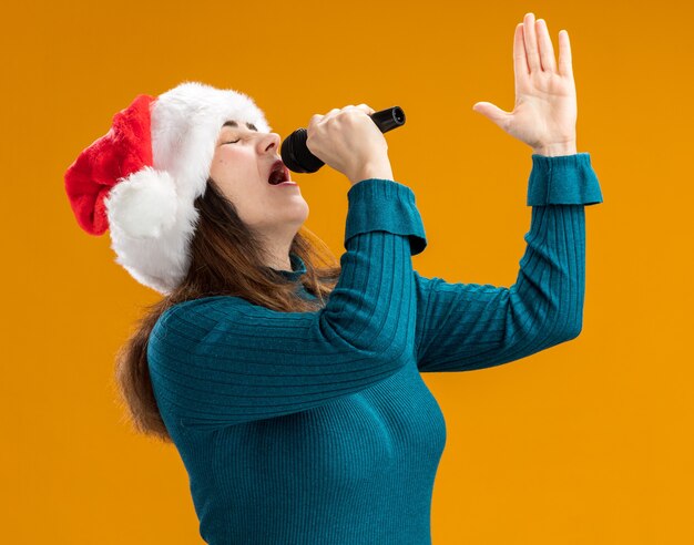 Caucasian woman with santa hat détient micro faisant semblant de chanter isolé sur fond orange avec copie espace