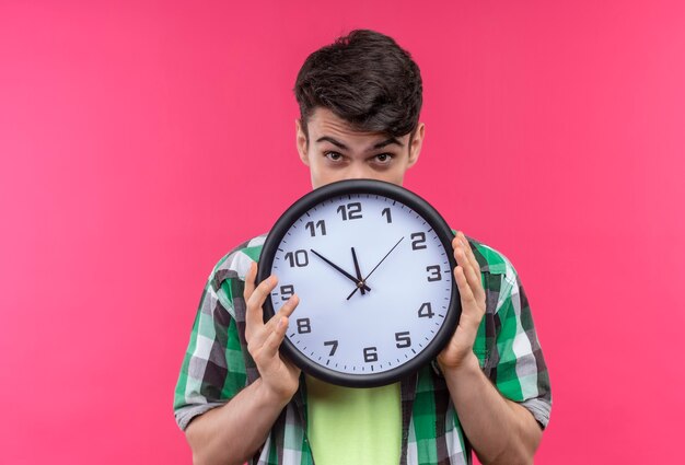 Caucasian young man wearing green shirt bouche couverte avec horloge murale sur mur rose isolé