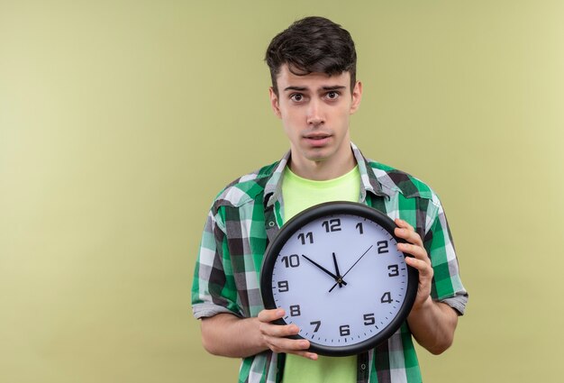 Caucasian young man wearing green shirt holding horloge murale sur mur vert isolé