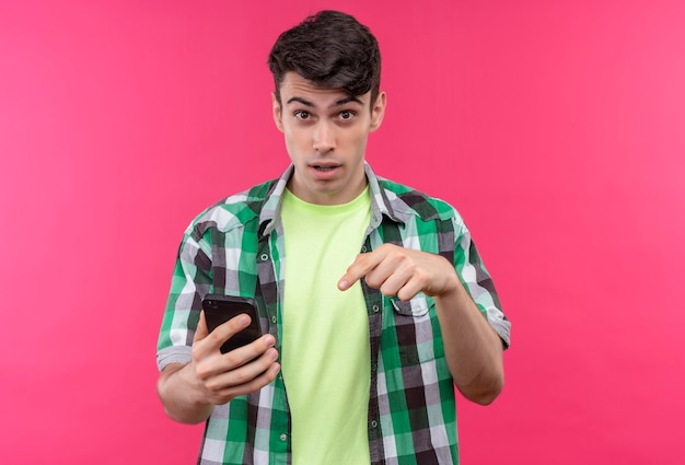 Caucasian young man wearing green shirt points at phone dans sa main sur un mur rose isolé