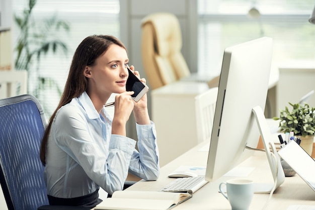 Caucasien employé féminin assis au bureau en face de l'écran et parlant sur smartphone