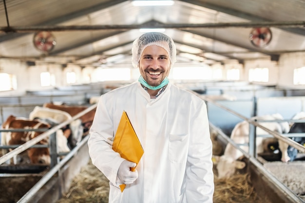 Caucasien souriant vétérinaire mal rasé en uniforme de protection blanc debout dans la grange avec dossier avec les résultats des tests sous l'aisselle.