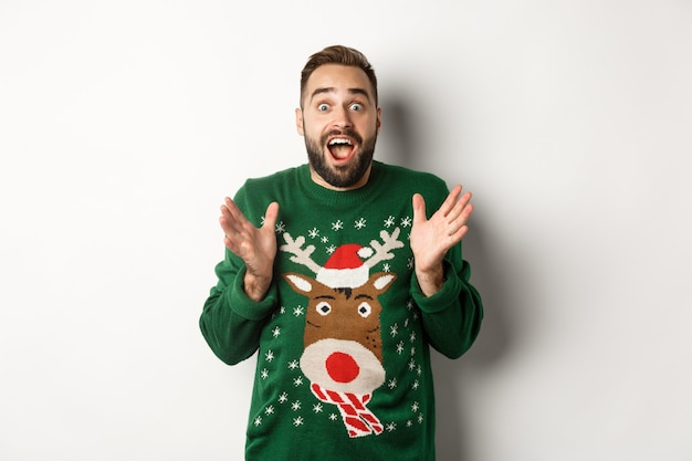 Célébration du nouvel an et concept de vacances d'hiver. Heureux et surpris barbu qui a l'air étonné, attrape quelque chose, debout dans un drôle de pull de Noël, fond blanc.