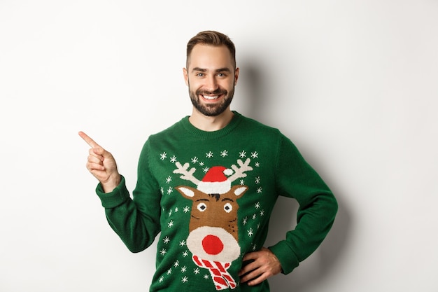 Célébration du nouvel an et concept de vacances d'hiver. Homme confiant et heureux avec barbe, portant un pull de Noël, pointant sur la bannière du coin supérieur gauche, fond blanc.