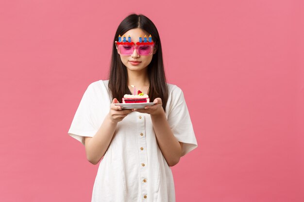 Célébration, fêtes et concept amusant. Fille d'anniversaire mignonne rêveuse dans des lunettes de soleil drôles tenant un gâteau de jour et regardant une bougie réfléchie, faisant un vœu, debout sur fond rose.