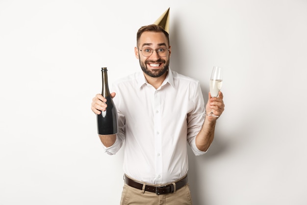 Célébration et vacances. Joyeux anniversaire gars appréciant la fête du b-day, portant un chapeau cône drôle et buvant du champagne