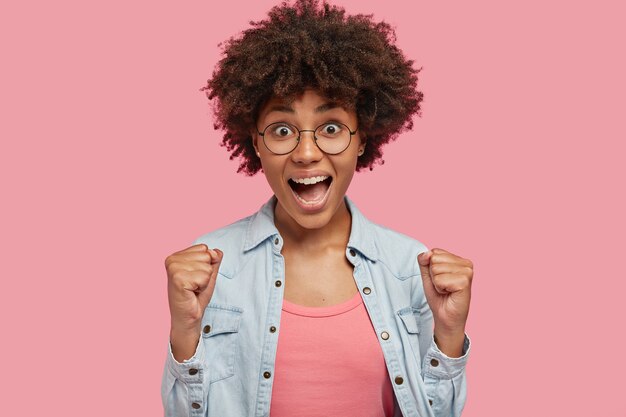 Célébrons mon succès! Jolie femme à la peau foncée et bouclée lève les poings fermés, se réjouit de triompher ou de gagner, porte des lunettes rondes et une veste en jean, se sent ravie et amusée, isolée sur un mur rose