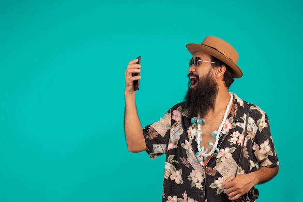 Celui d'un homme heureux à la longue barbe coiffé d'un chapeau, vêtu d'une chemise à rayures, tenant un téléphone sur un fond bleu.