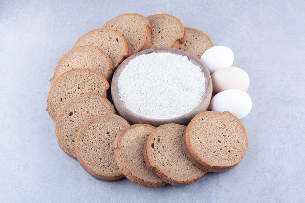 Cercle de tranches de pain noir et oeufs autour d'un bol de farine sur une surface en marbre