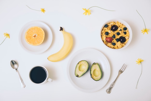 Céréales cornflakes; Avocat; banane; orange coupée en deux; café et fleurs sur fond blanc