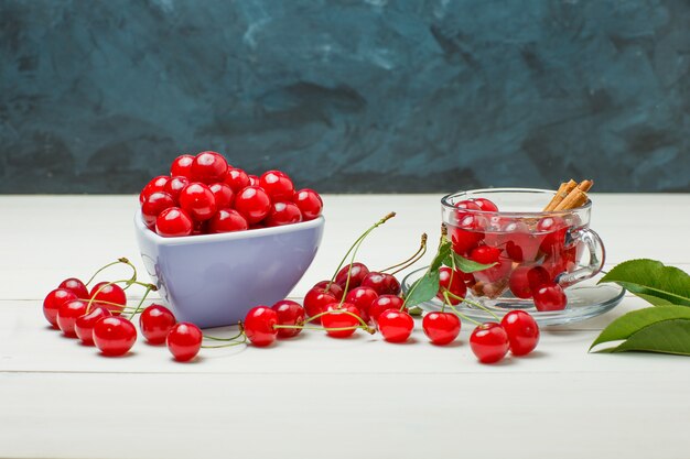 Cerises aux feuilles, épices dans un bol et mug sur bois et bleu foncé, vue latérale.