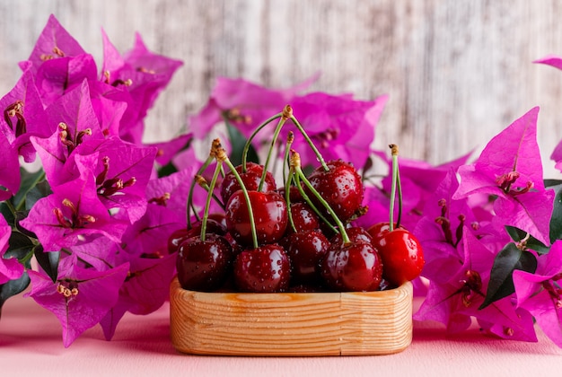 Cerises dans une assiette en bois avec des fleurs vue de côté sur une surface rose et grungy