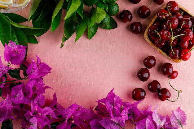 Cerises avec fleurs, citrons, feuilles, dans une assiette en bois sur une surface rose, vue du dessus