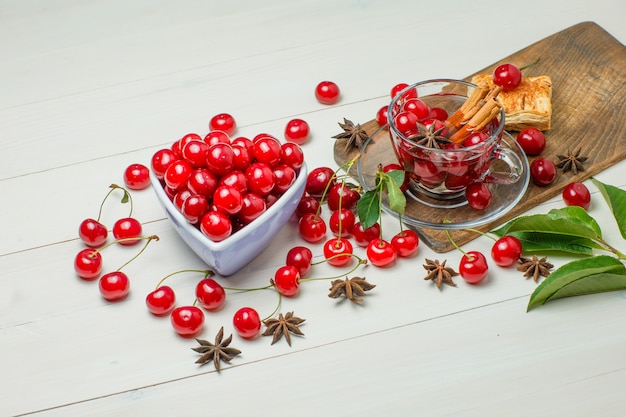 Cerises avec pâtisserie, feuilles, épices dans un bol et verre sur planche de bois et à découper, vue grand angle.