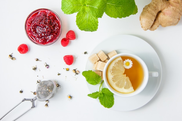 Certains une tasse de thé à la camomille avec des herbes, des framboises et de la confiture, des feuilles de menthe, du sucre sur la surface blanche, vue de dessus.