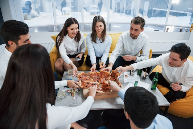 Photo gratuite chacun a sa propre tranche. manger de la pizza. célébration d'une transaction réussie. jeunes employés de bureau assis près de la table avec de l'alcool