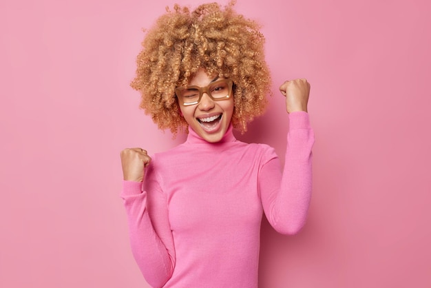 Chanceuse positive jeune femme aux cheveux bouclés rit et cligne de l'oeil fait un geste de triomphe porte des lunettes et poloneck célèbre le triomphe isolé sur fond rose Oui finalement j'ai gagné le succès