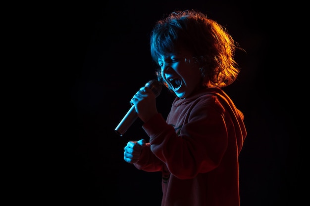 Chanter comme une célébrité, rockstar. Portrait de garçon caucasien sur fond sombre de studio en néon. Beau modèle bouclé. Concept d'émotions humaines, expression faciale, ventes, publicité, musique, passe-temps, rêve.