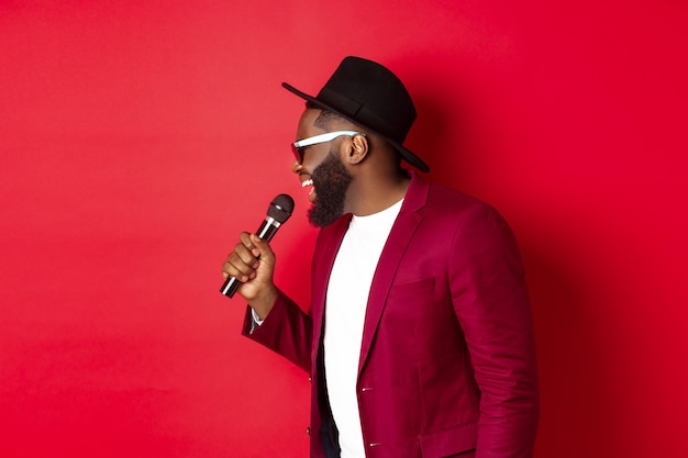 Photo gratuite chanteur noir passionné se produisant sur fond rouge, chantant dans un microphone, portant une tenue de fête, debout sur fond rouge.